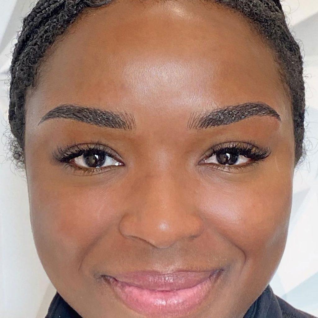 Close-up of a smiling woman with defined, softly shaded brows that darken toward the tails, wearing lashes and a dark top.